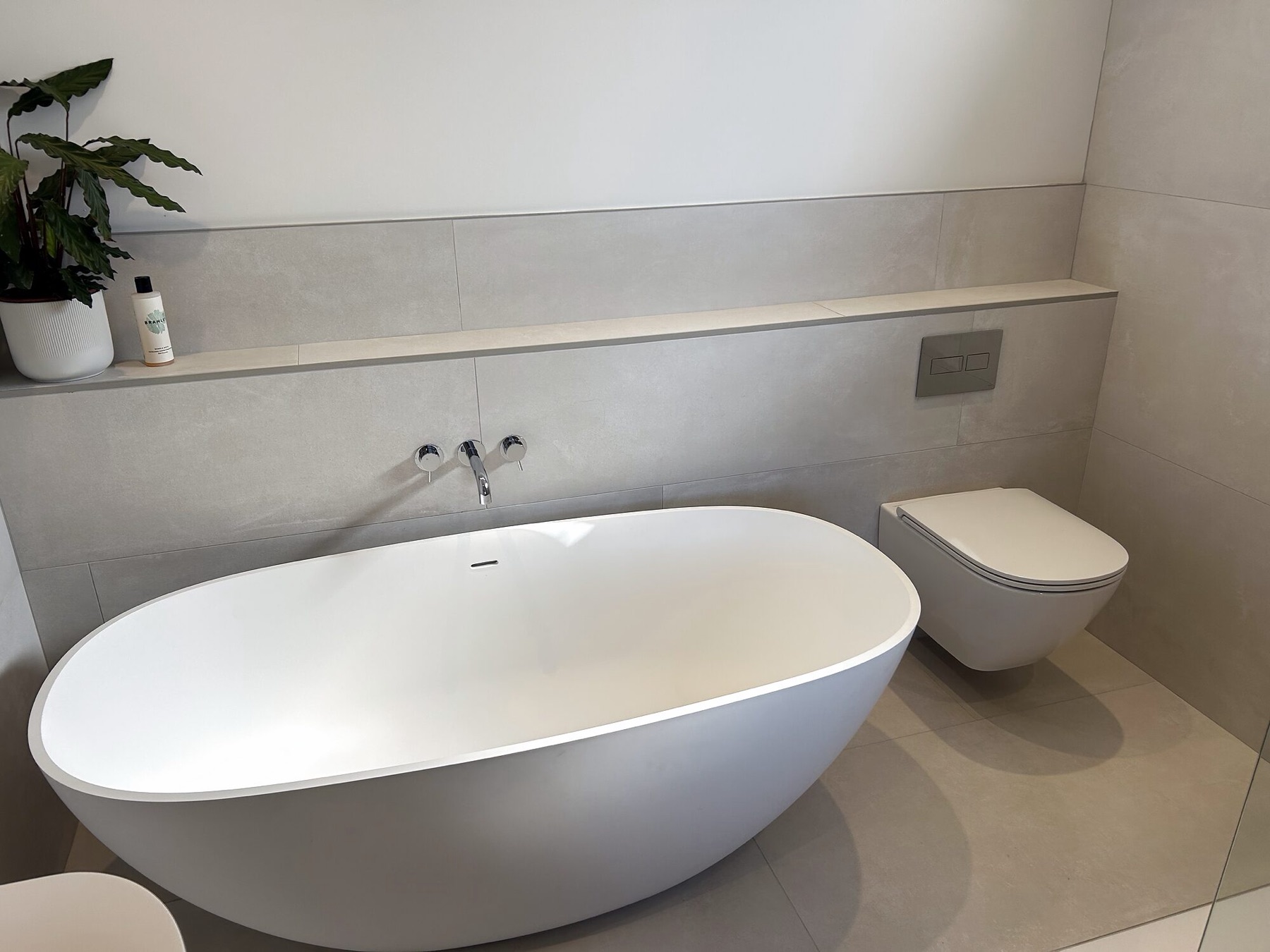 Freestanding bath against pale tiles, with a wall-hung toilet and concealed cistern.